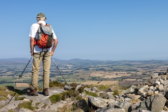 A walker stands on top of a rocky outcrop, holding walking poles and a back pack on his back - he looks out over the expanse of fields in front of him.