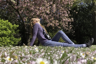 A beautiful young woman sits on the grass in a park, with daisies in the foreground and blossoming trees behind. She has her head thrown back and her eyes closed, enjoying the peace of nature