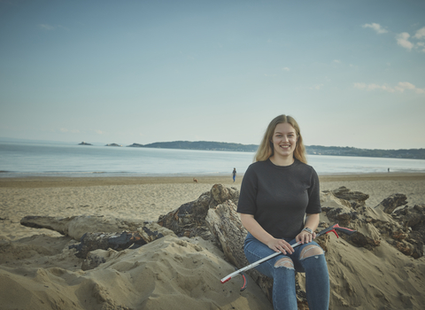 A woman sits on a beach with the sea in the background, holding a litter picker, smiling at the camera
