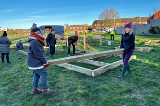 Two women carry a wooden plank as others build raised beds in the background. 