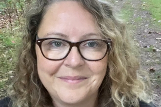 Miriam is in a woodland, her head and shoulders and visible and she is smiling at the camera