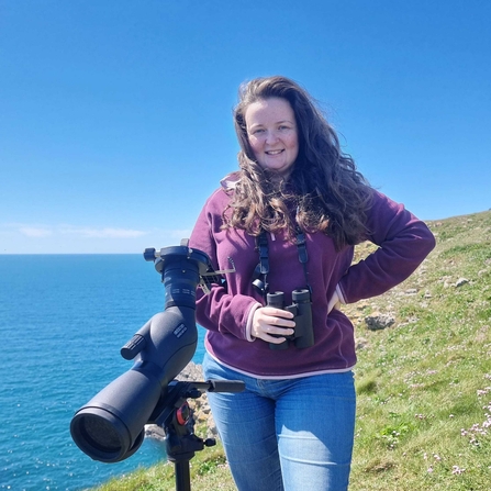 Sarah Cunningham stands on a sea cliff with her binoculars. 