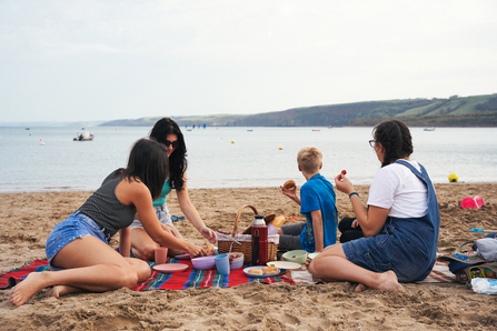 Four people sit on the beach, eating a picnic, gazing out to sea