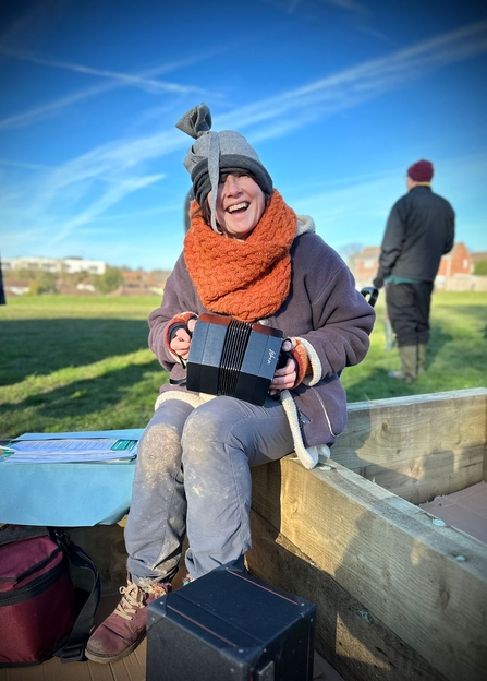 A woman in winter clothes plays a concertina outside with blue sky and grass in the background. 