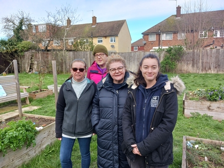 Four people stand in a community garden with raised beds to either side. 