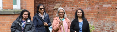 Four women smiling and holding gardening tools with red brick wall background. 