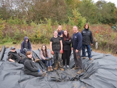 A group of young people stand in a recently dug out pond before it is filled with water