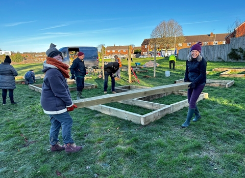 Two women carry a wooden plank as others build raised beds in the background. 